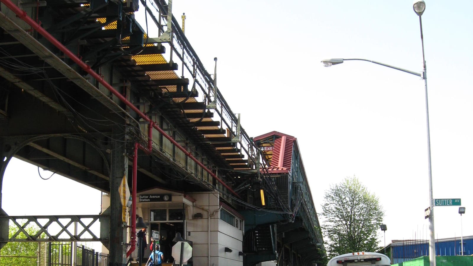 Photograph from street level of an elevated subway structure with a red pipe and steel beams, a small station house signed Sutter Avenue Station with an L symbol, a red-roofed stair enclosure, a tall street light, a green street sign reading Sutter Av, a tree and the top of a white bus, with two people seen from behind at the station door