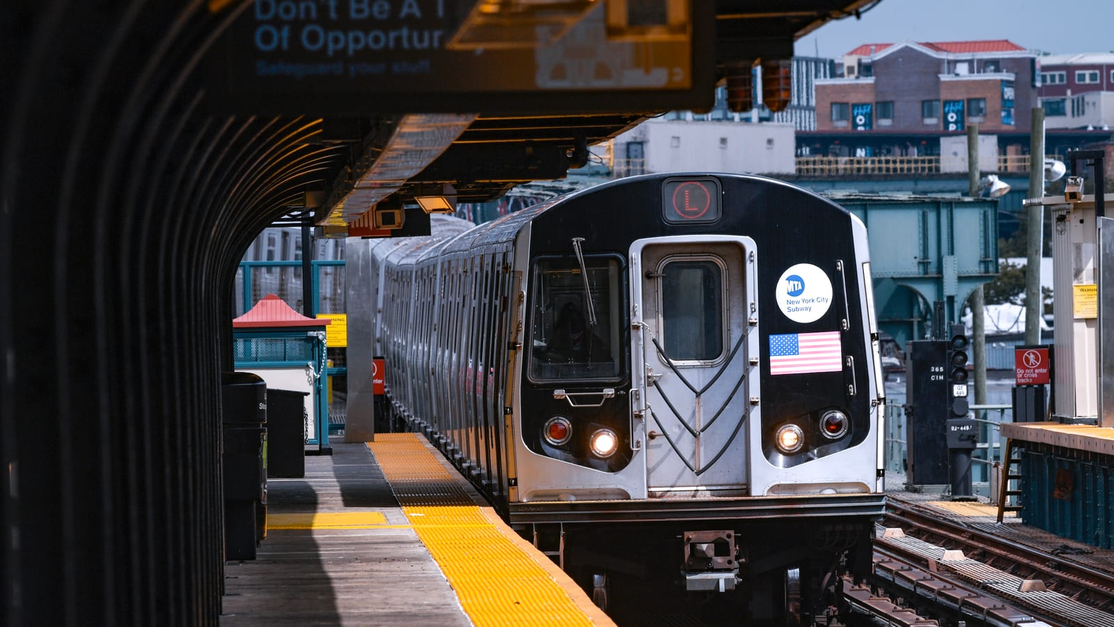 Photograph of a silver L train with an MTA New York City Subway badge and an American flag decal pulling into an elevated station, with a dark arched platform canopy at left, a yellow platform edge, signal equipment at right and low brick buildings behind