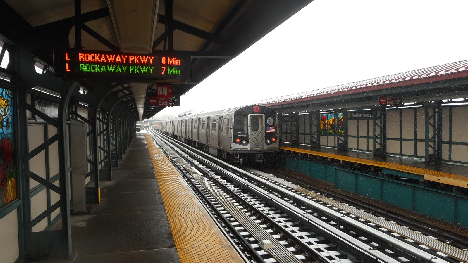 Photograph of an elevated subway platform in light snow, with a dark canopy, an orange and green sign reading L Rockaway Pkwy 0 Min and 7 Min, a yellow platform edge strip, a silver L train on the far track beside a second platform with a red roof, stained-glass panels and a Sutter Avenue sign