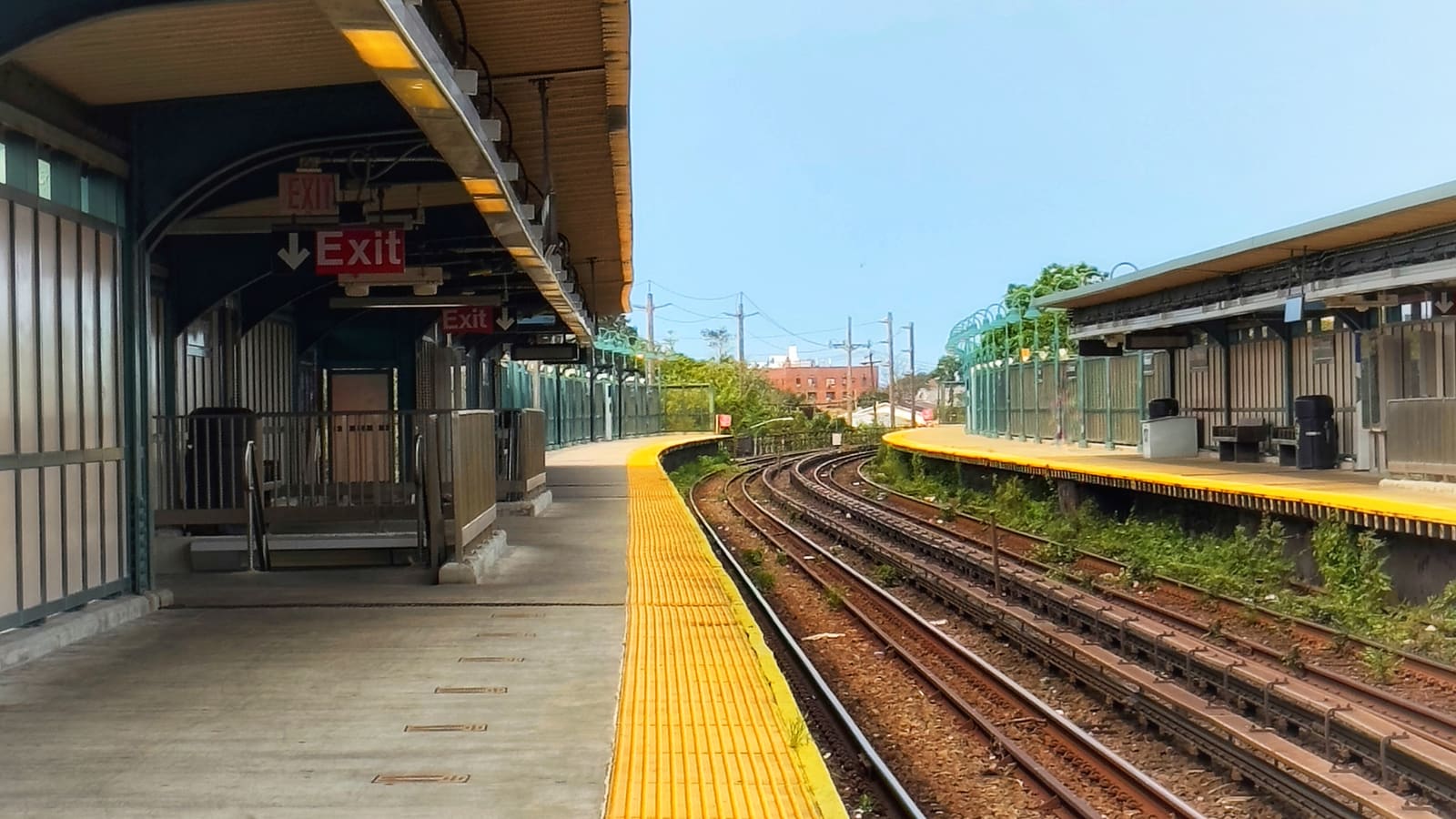 Photograph of an empty elevated subway platform with a yellow edge strip, a covered area with red Exit signs and a stairway at left, curving tracks in the middle and a second covered platform with benches and trash cans at right, trees and a red brick building in the distance, under a pale blue sky