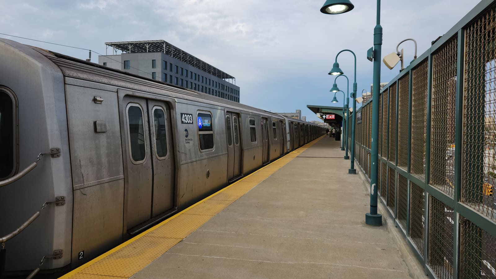 Photograph of a silver subway train with an A sign on its side stopped at an elevated open-air platform with a yellow edge strip, green lamp posts and a green mesh fence at right, a red Exit sign under a canopy in the distance, and a gray building with solar panels on its roof behind the train, under a cloudy sky