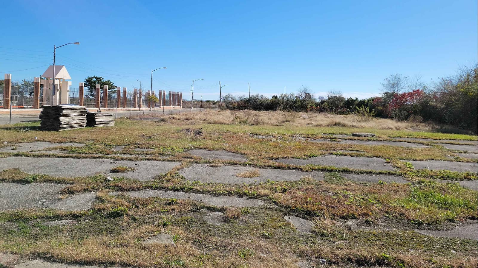 Photograph across a vacant lot of cracked pavement patches and grass, with two stacks of wooden boards at left, a row of brick posts with a fence and a small white gatehouse beyond, street lights, and low shrubs with red leaves at right, under a clear blue sky