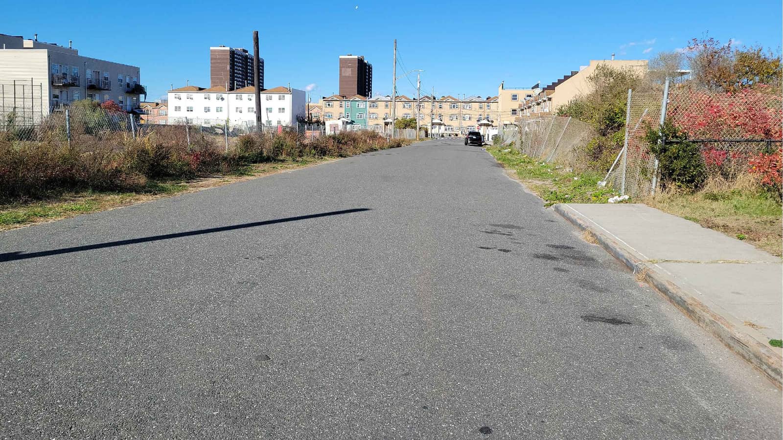 Photograph down an empty two-lane street with a sidewalk at right, chain-link fences and red-leaved shrubs on both sides, rows of three-story attached houses and a white apartment building ahead, and two tall brown towers in the distance, under a clear blue sky