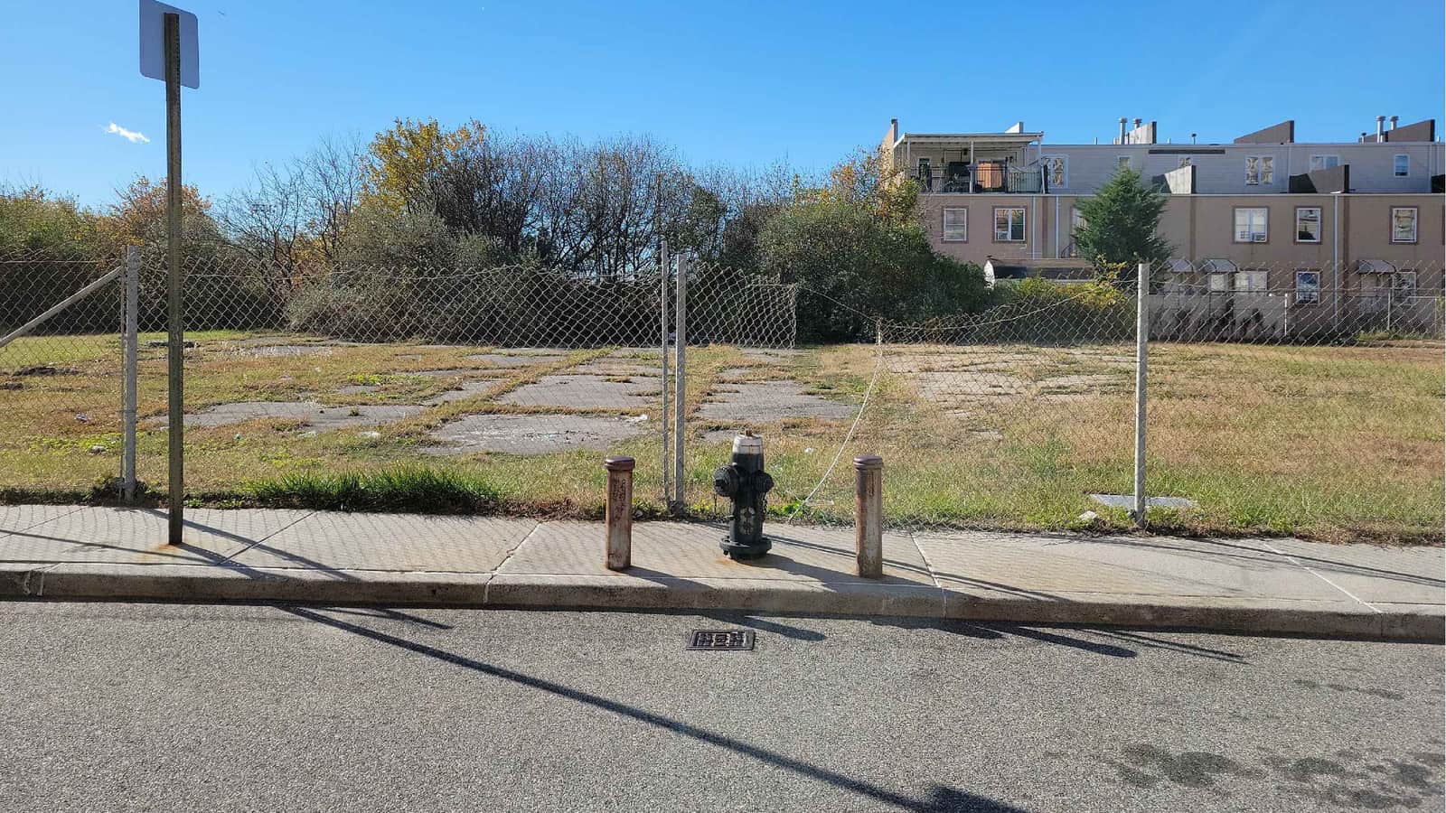 Photograph of a vacant lot behind a chain-link fence, with patches of broken pavement and grass, a sidewalk with a black fire hydrant between two short metal posts and a sign pole in front, trees at left and a row of three-story beige houses at right, under a clear blue sky