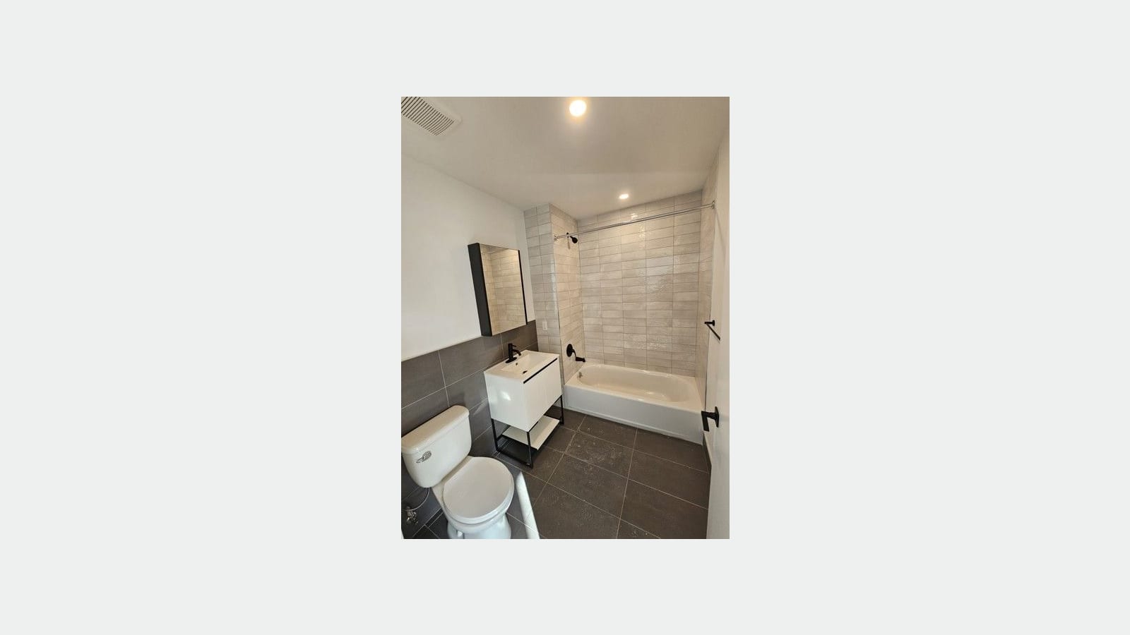 Photograph of a bathroom with a white tub and tiled walls, a curved shower rod, a white vanity on a black metal frame with a black faucet, a mirrored cabinet, a white toilet and dark gray floor tiles