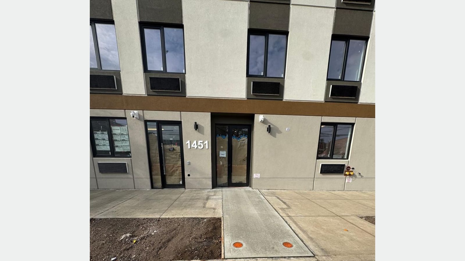 Photograph of the building's ground floor: two glass doors with the number 1451 between them, wall lights, security cameras and a window with papers taped inside; above, windows with air-conditioner grilles under a gold band; a new concrete sidewalk with a bare tree pit in front