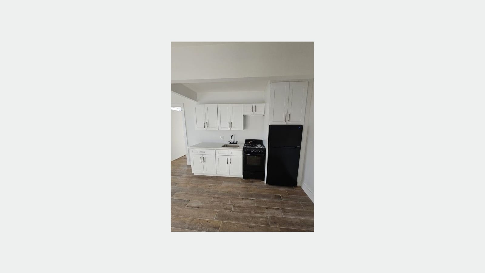 Photograph of a kitchen along one wall with white shaker cabinets, a small sink with a black faucet, a black range with electric coil burners, a black refrigerator under a tall cabinet and a wood-look tile floor