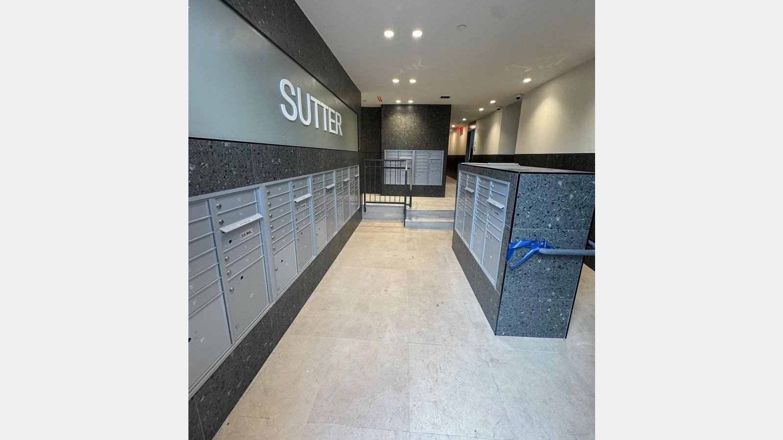 Photograph of a lobby with a light stone-look tile floor, rows of gray metal mailboxes set into speckled dark gray stone walls, the word SUTTER in raised white letters on a gray-green panel, recessed ceiling lights and a short stair with a railing at the back