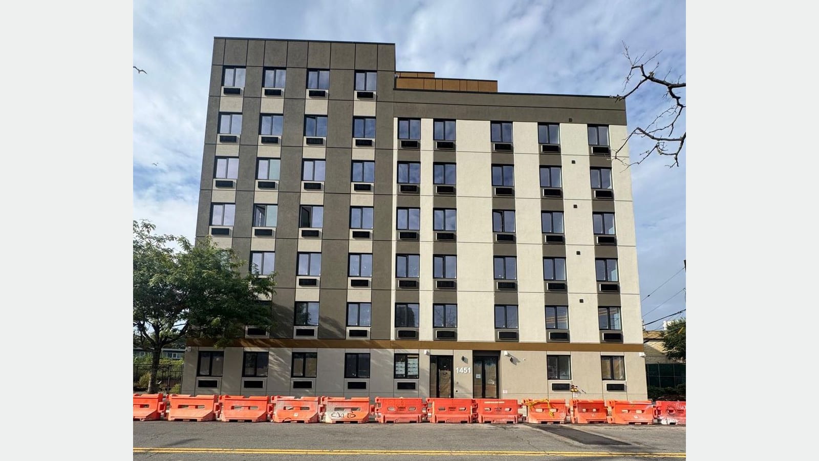 Photograph of a seven-story apartment building with dark gray and cream panels, rows of windows with air-conditioner sleeves under them, a gold band above the ground floor and the number 1451 beside two glass entry doors; a line of orange plastic traffic barriers runs along the curb, with a tree at left under a partly cloudy sky