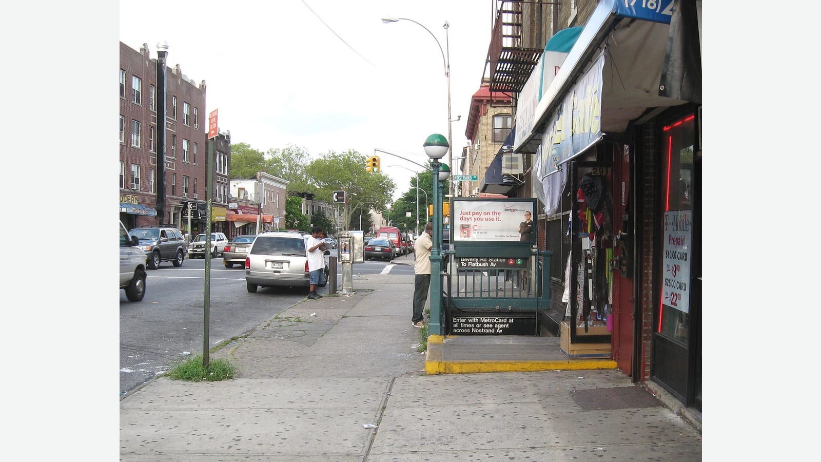 Photograph of a sidewalk on Nostrand Avenue with a green-railed subway stair entrance signed Beverly Rd Station, shop awnings and a fire escape on the right, parked cars and brick apartment buildings across the street, and a few people in the distance