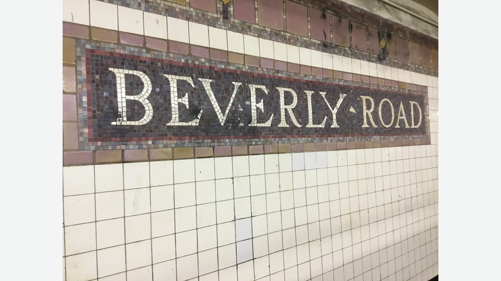 Photograph of a dark mosaic name tablet reading BEVERLY ROAD in white letters with a red border, set in a white tiled subway station wall