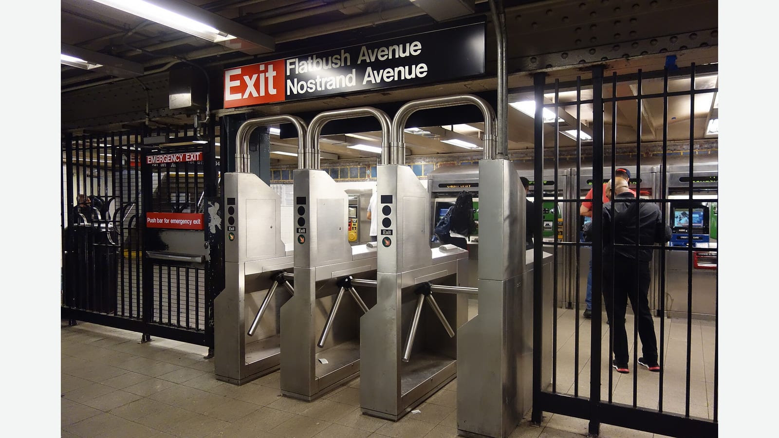 Photograph of a row of steel subway turnstiles under a black sign reading Exit Flatbush Avenue Nostrand Avenue, with a barred emergency gate on the left and riders seen from behind at the vending machines on the right