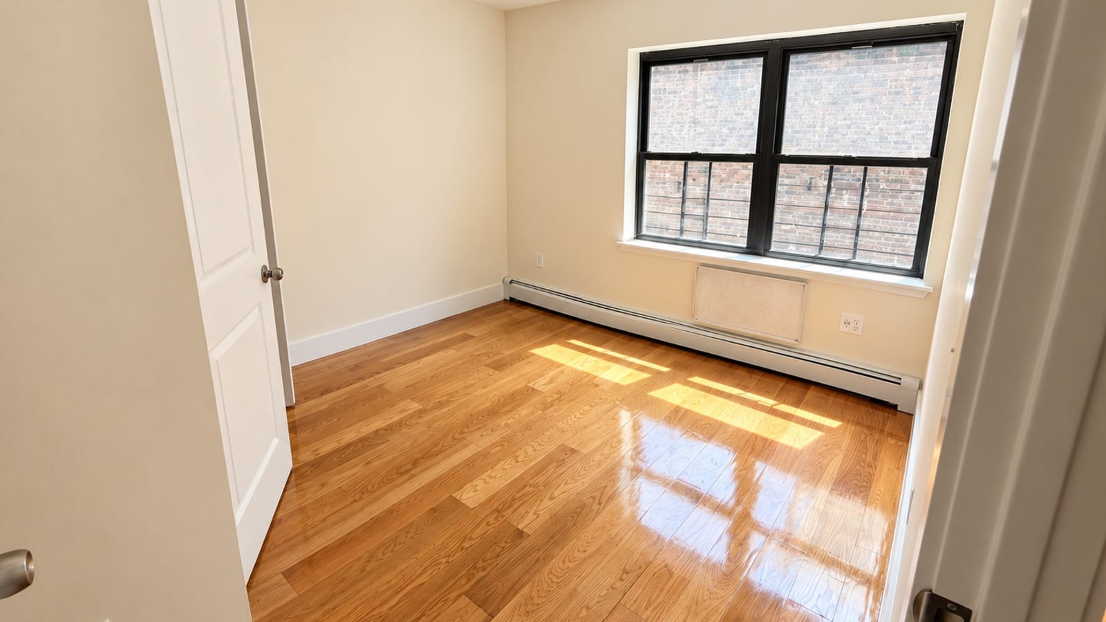 Photograph of an empty room seen through an open doorway, with a glossy wood floor lit by sun, cream walls, a white panel door at left, a black-framed double window with window guards facing a brick wall, a covered air-conditioner sleeve and a baseboard heater