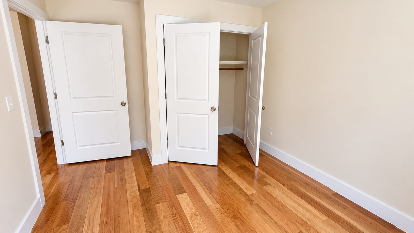Photograph of an empty room with a wood floor, cream walls and white baseboards, an open white panel door to a hallway at left, and a closet with two open white panel doors showing a shelf and a hanging rod