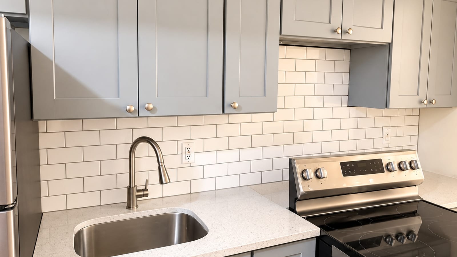 Photograph of a kitchen wall with gray shaker upper cabinets with round knobs, white subway tile, a white speckled counter with an undermount stainless steel sink and a curved faucet, and a stainless steel range with knobs and a smooth black cooktop at right