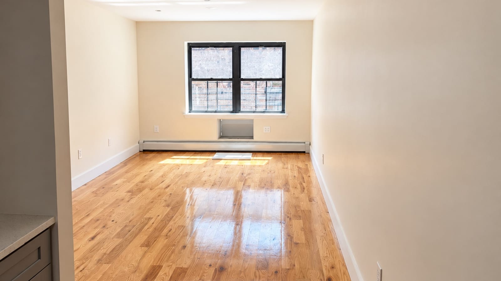 Photograph of an empty, sunlit room with a glossy light wood floor, cream walls, white baseboards, a black-framed double window looking onto a brick wall, an air-conditioner sleeve and a long baseboard heater under the window, and the edge of a gray kitchen cabinet with a white counter at lower left