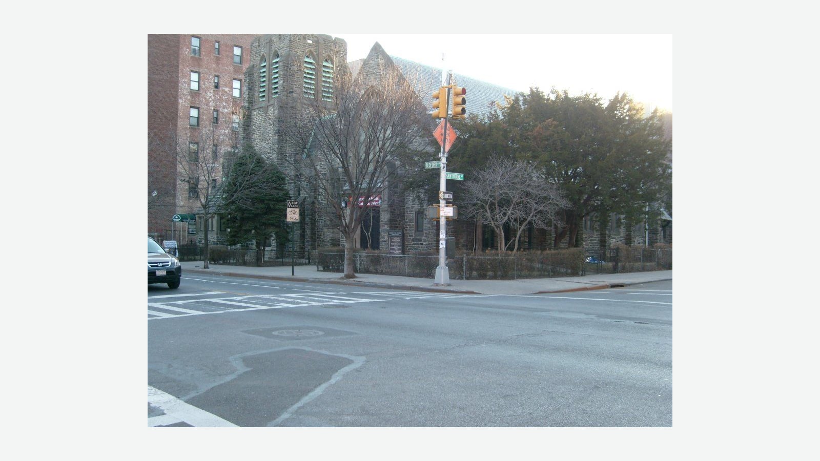 Photograph of a street corner with green street signs reading Bedford and Hawthorne on a pole with traffic lights and an orange diamond sign, a gray stone church with a tower behind bare trees and an iron fence, a tall red brick apartment building at left, a bike lane sign, a parked car at the left edge and an empty crosswalk in the foreground, shown at its original size on a plain light background