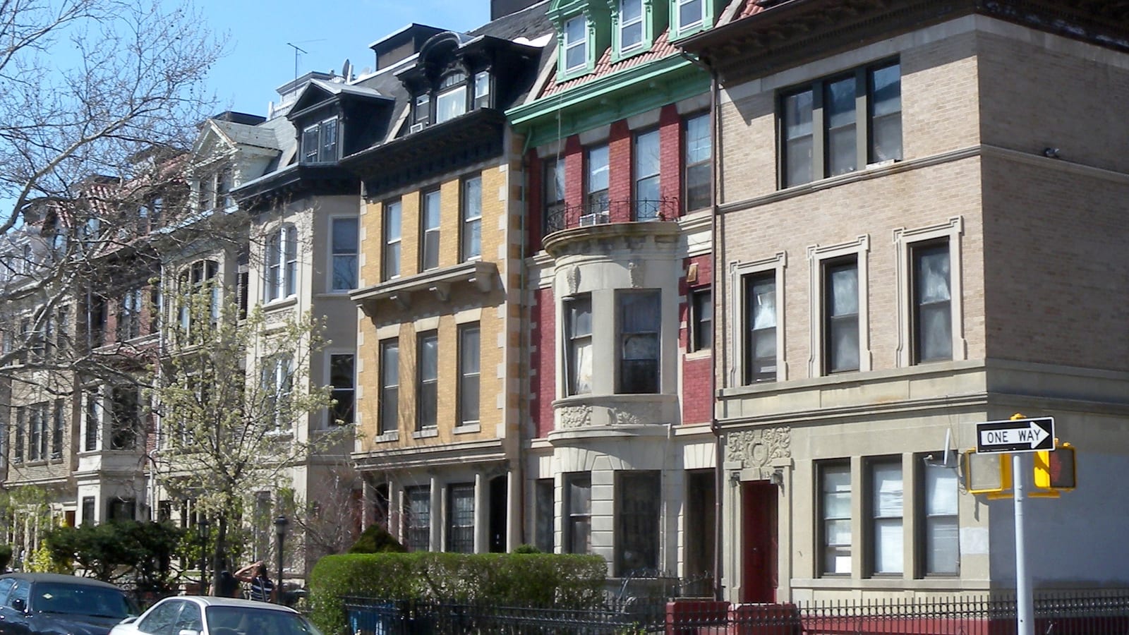 Photograph of a row of three- and four-story attached houses in yellow, red and tan brick with slate mansard roofs, green and black dormers, bay windows and an iron fence with a hedge, two parked cars at lower left, a one-way sign at right and a clear blue sky