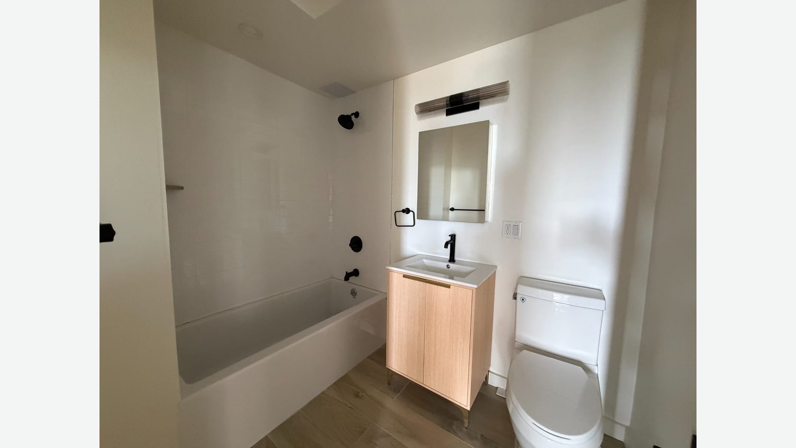 Photograph of a bathroom with a white tub and white tiled walls, black shower and tub fixtures, a light wood vanity with a white sink and black faucet, a mirrored cabinet under a black light bar, a black towel ring and a white toilet
