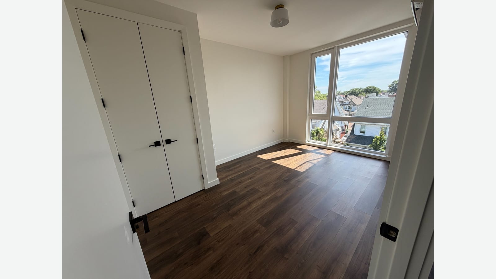 Photograph of an empty bedroom with white double closet doors with black handles, tall corner windows looking over nearby houses and trees, a ceiling light and dark plank flooring with sunlight on it