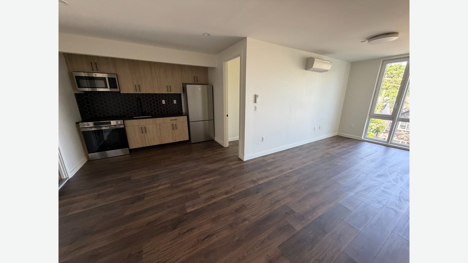 Photograph of an open kitchen and living area with light wood upper and lower cabinets, a black hexagon tile backsplash, a microwave over a stainless range, a stainless refrigerator, a white wall unit air conditioner, white walls, a corner window and dark plank flooring
