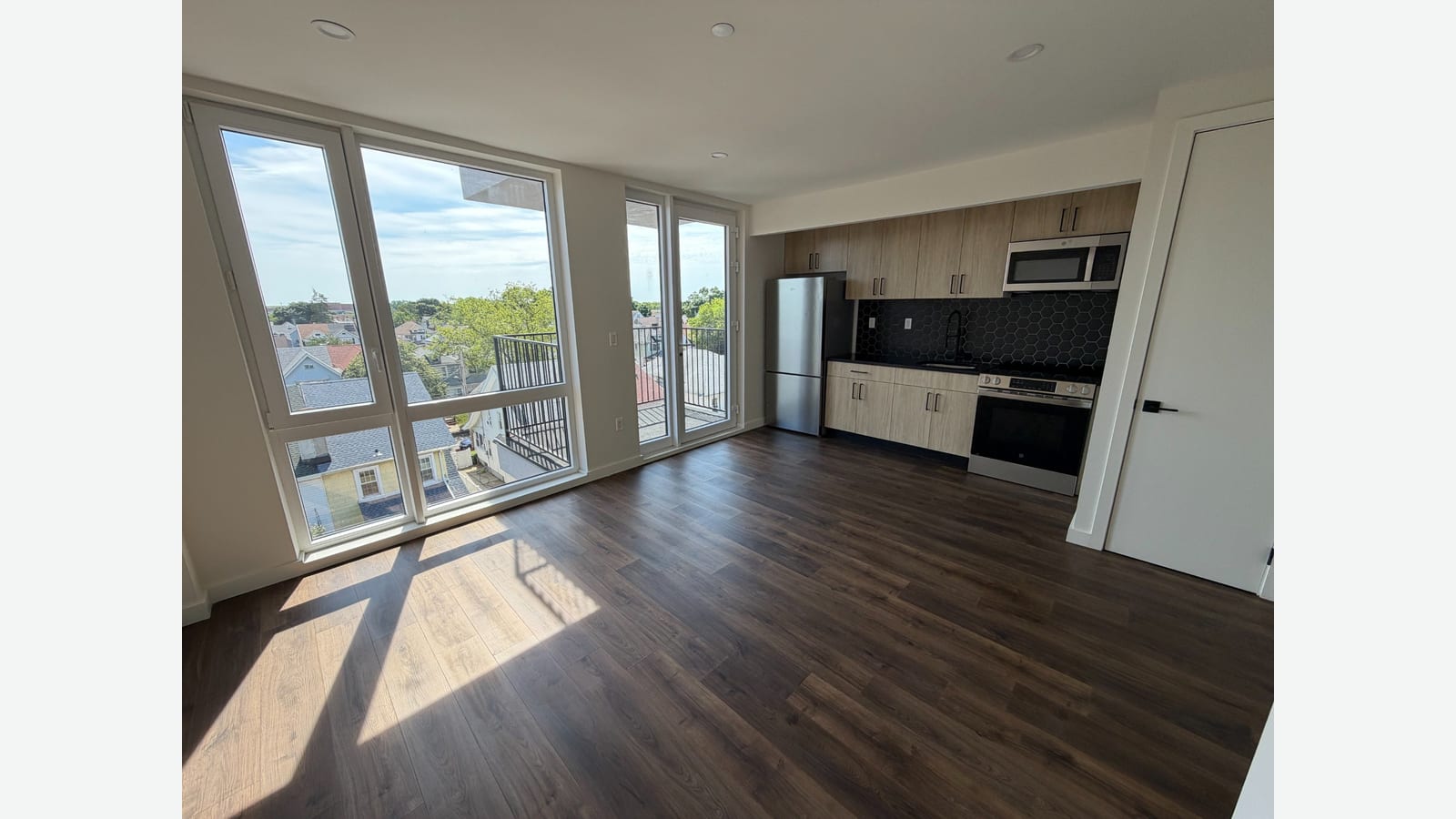 Photograph of an empty living room with dark wood-look plank flooring, tall corner windows looking over low rooftops and trees, a glass door to a small balcony with a black railing, and a kitchen along the far wall with light wood cabinets, a black tile backsplash, a stainless refrigerator, microwave and range