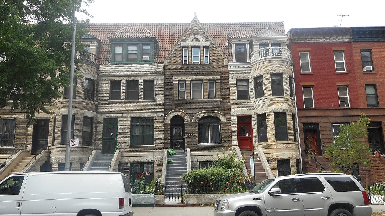 Photograph of a row of three-story stone row houses with tiled roofs, a pointed central gable, rounded bay windows and stoops, next to a red brick row house, with a white van and a silver SUV parked in front