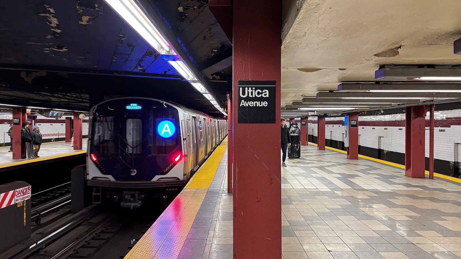 Photograph of an underground subway platform with red columns and a black sign reading Utica Avenue, an A train with a blue A sign pulling in on the left, and a few distant riders on the tiled platform
