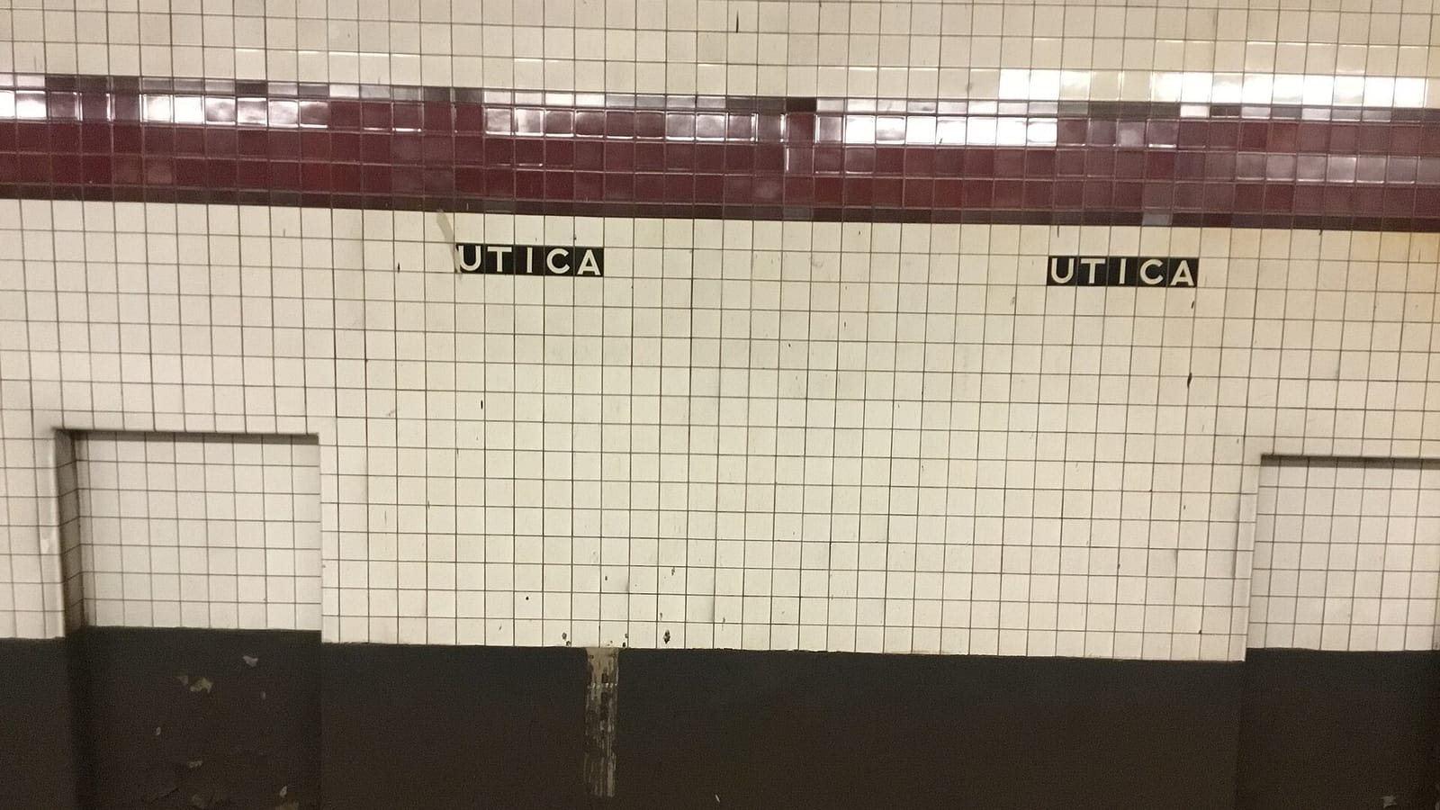 Photograph of a white tiled subway wall with a dark red tile band and two small black name tablets reading UTICA, above a dark lower wall