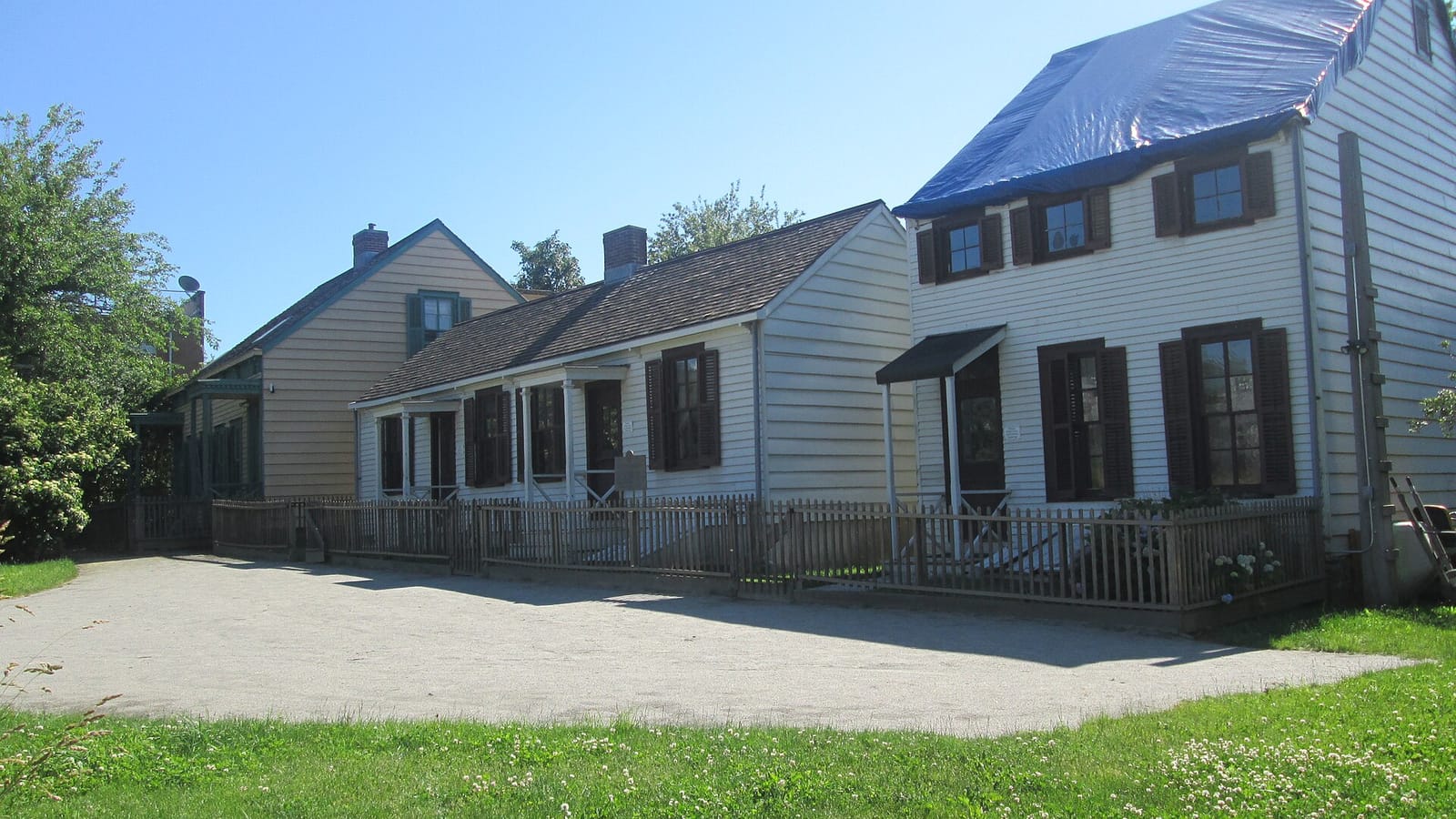 Photograph of three small white and pale yellow wooden houses with dark shutters behind a low picket fence, one with a blue tarp over its roof, beside a gravel path and a green lawn under a blue sky