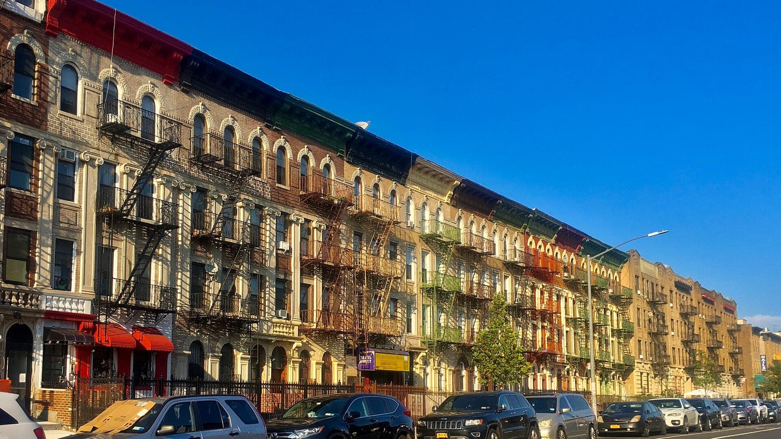 Photograph of a long row of five-story brick and painted apartment buildings with arched windows, black fire escapes and colorful cornices, cars parked along the curb and a small shop sign at street level, under a clear blue sky