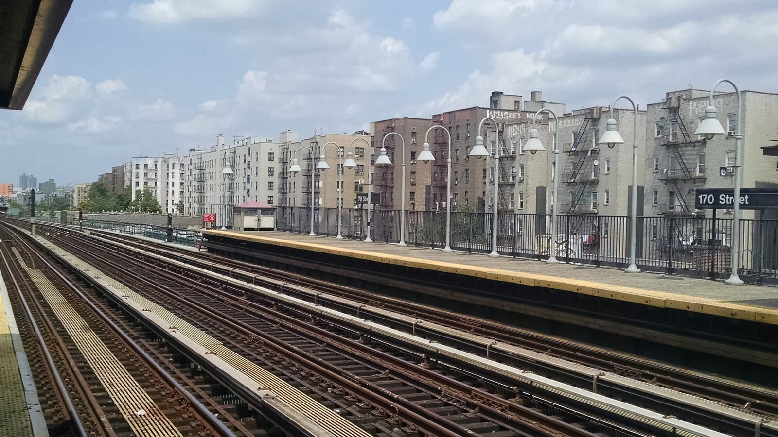 Photograph across the tracks of an elevated subway station, with an open platform lined with white lamp posts, a black sign reading 170 Street on the right and apartment buildings with fire escapes behind under a partly cloudy sky