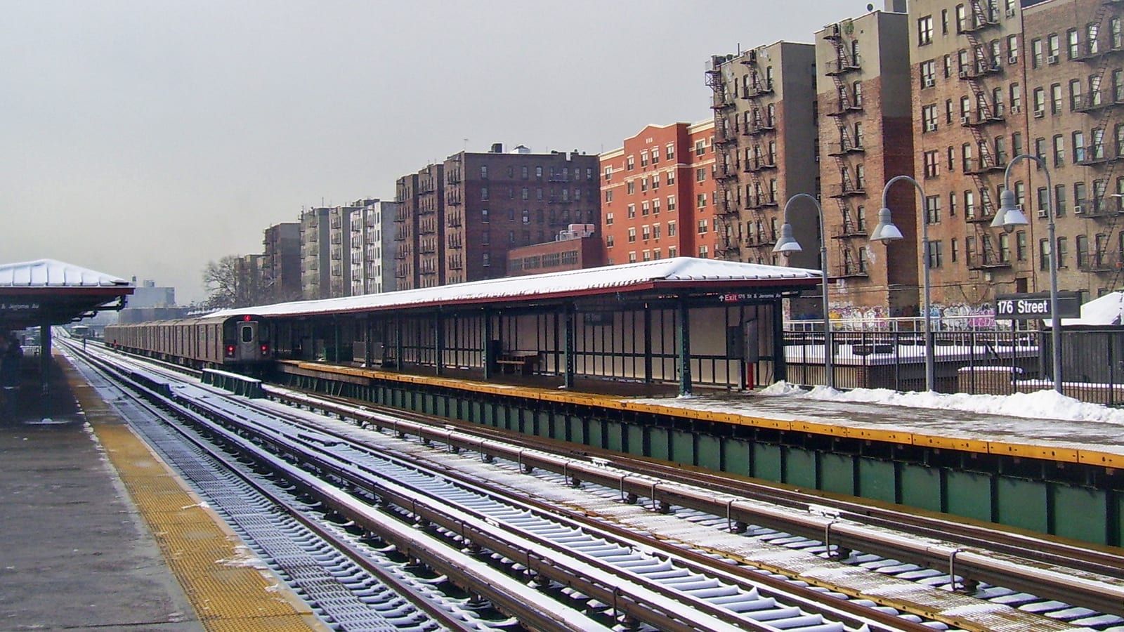 Photograph of an elevated subway station in snow, with a long platform shelter, a sign reading 176 Street on the right, a train leaving to the left and a row of brick apartment buildings with fire escapes behind