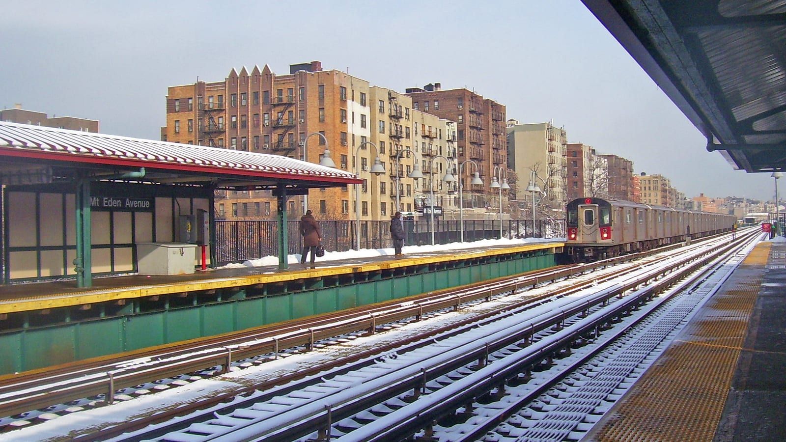 Photograph of an elevated subway station in snow, with a red-roofed platform shelter and a sign reading Mt Eden Avenue on the left, two people waiting far off, a silver train arriving on the right track and tan and brown apartment buildings behind