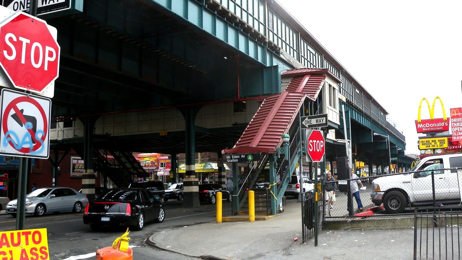 Photograph of a covered red stair climbing to an elevated green steel subway structure, with a sign reading Mt Eden Av Station and a green 4 symbol, stop signs and one-way signs, parked cars under the tracks, two men on the sidewalk and a McDonald's sign on the right