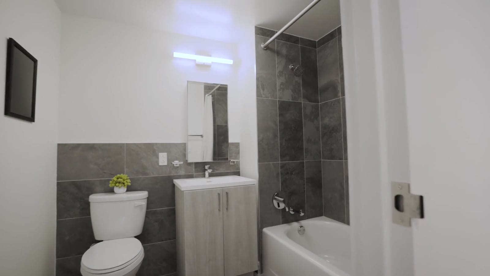 Photograph of a bathroom with dark grey stone-look tile on the lower walls and around a white bathtub, a white toilet, a small vanity with a pale wood-look cabinet, a mirrored cabinet and a bar light above it