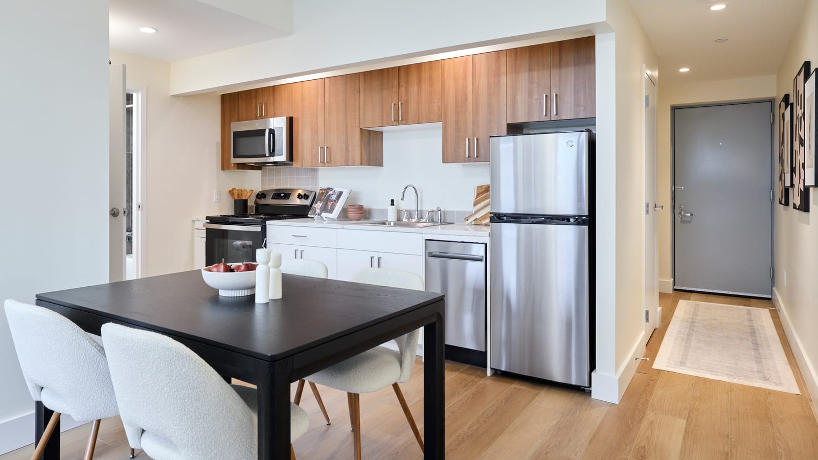 Photograph of an open kitchen with wood-tone upper cabinets, white lower cabinets, a stainless-steel refrigerator, dishwasher, range and microwave, a black dining table with white chairs in the foreground and a hallway leading to a grey entry door