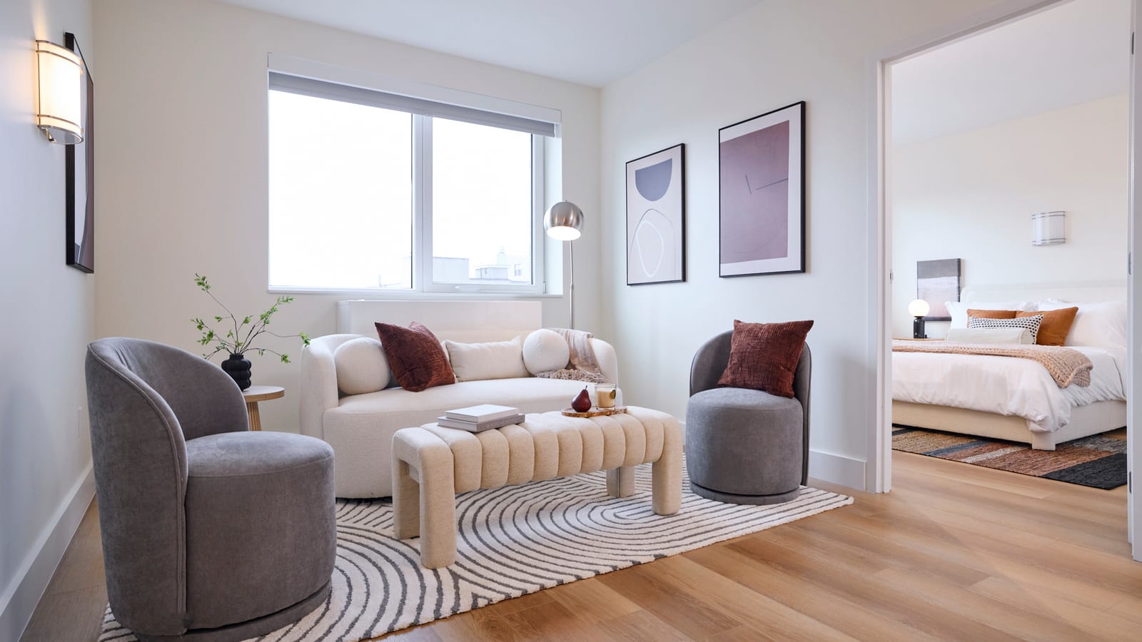 Photograph of a furnished living room with pale walls, wood-look plank flooring, a two-pane window with a roller shade, a white sofa, two grey armchairs, an upholstered bench on a striped rug, two framed prints and a doorway to a bedroom with a made bed
