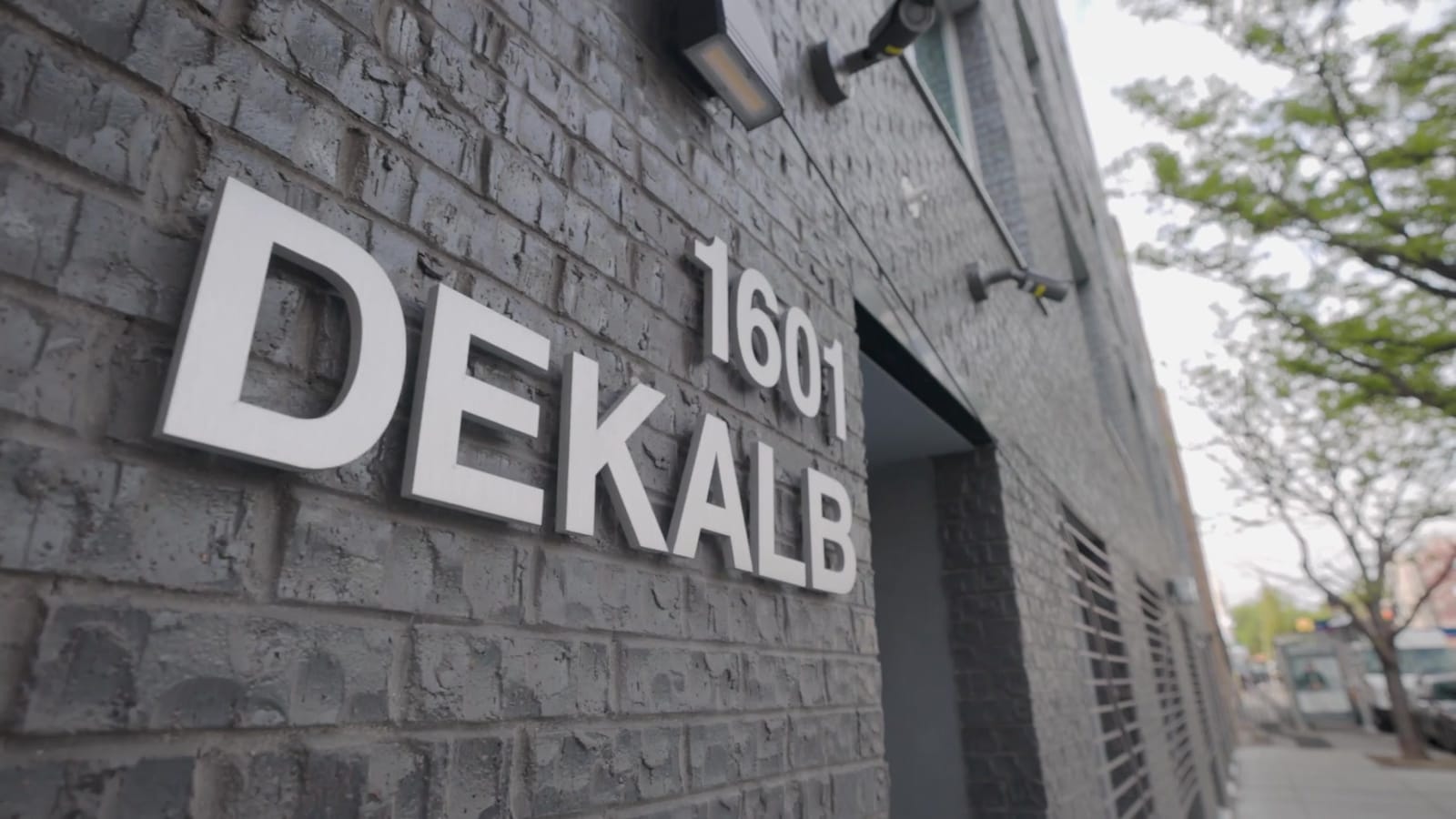 Close photograph of metal letters reading 1601 DEKALB mounted on a dark grey brick wall beside a recessed entrance, with a light fixture and security cameras above and street trees along the sidewalk