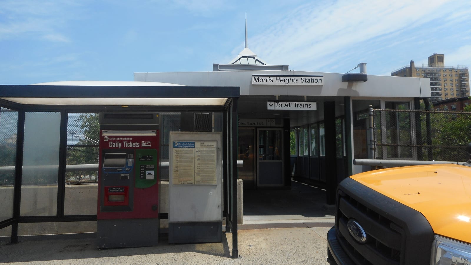 Photograph of a small station entrance building with a sign reading Morris Heights Station and a To All Trains sign, a red Metro-North ticket machine and a schedule board under a glass shelter, and the front of a yellow pickup truck at right