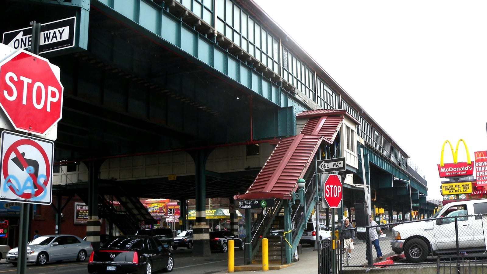 Photograph of a street corner under an elevated subway line, with a red-roofed stair rising to the Mt Eden Av station, stop and one-way signs at left, parked cars, a McDonald's sign at right and two distant pedestrians
