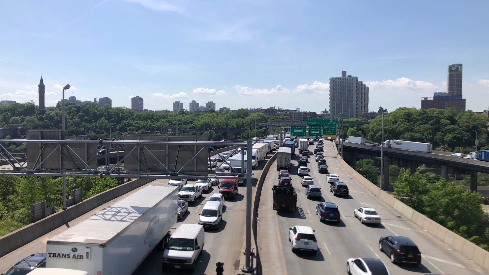 Photograph looking down from an overpass at a crowded multi-lane highway with trucks and cars, overhead green signs, a parallel elevated roadway at right, and wooded slopes with apartment towers and a stone water tower on the skyline under a blue sky