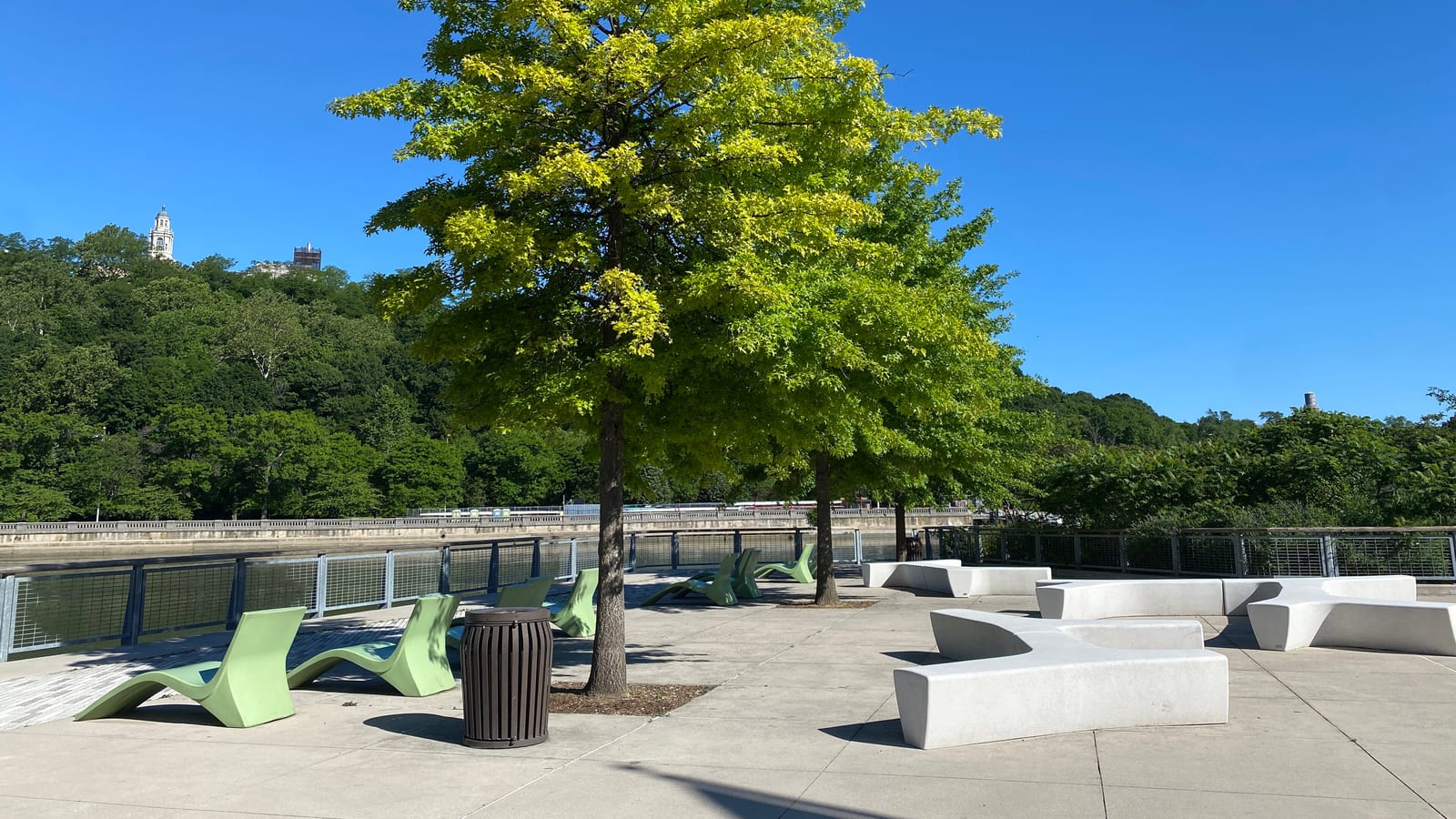 Photograph of a riverside plaza in summer with green lounge chairs, white stone benches, a trash bin and a young tree with yellow-green leaves in front of a metal railing, a river behind it and a wooded bluff with a tower on the far shore under a clear blue sky
