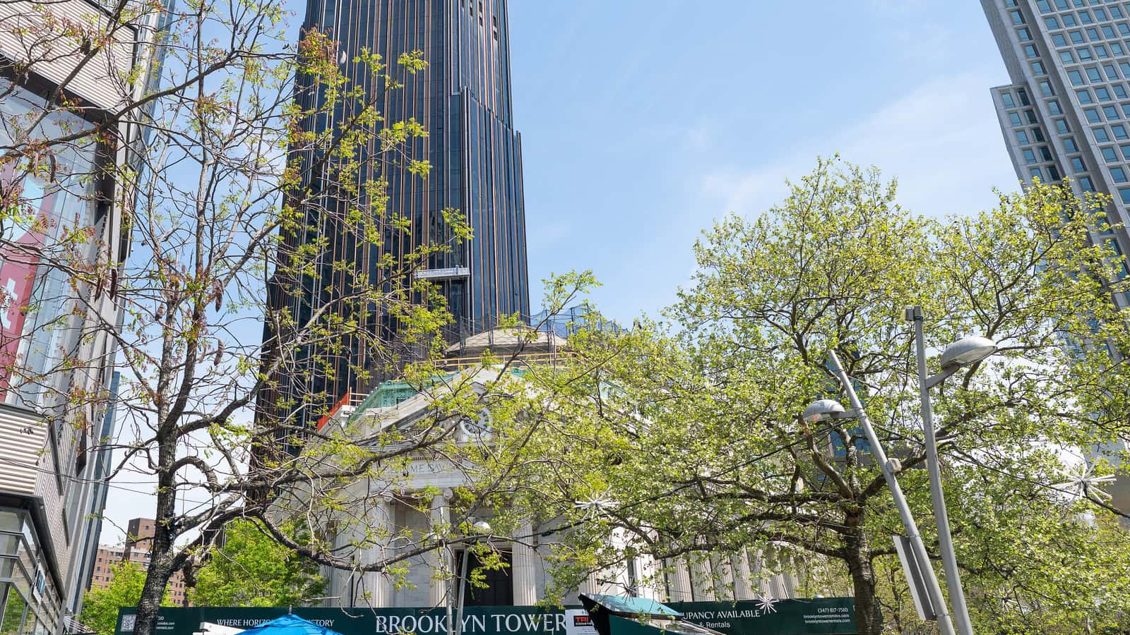 Photograph of a tall dark tower rising behind a domed classical bank building with columns, framed by trees in spring leaf, with a glass tower at right, blue umbrellas and a green construction fence reading Brooklyn Tower at the bottom