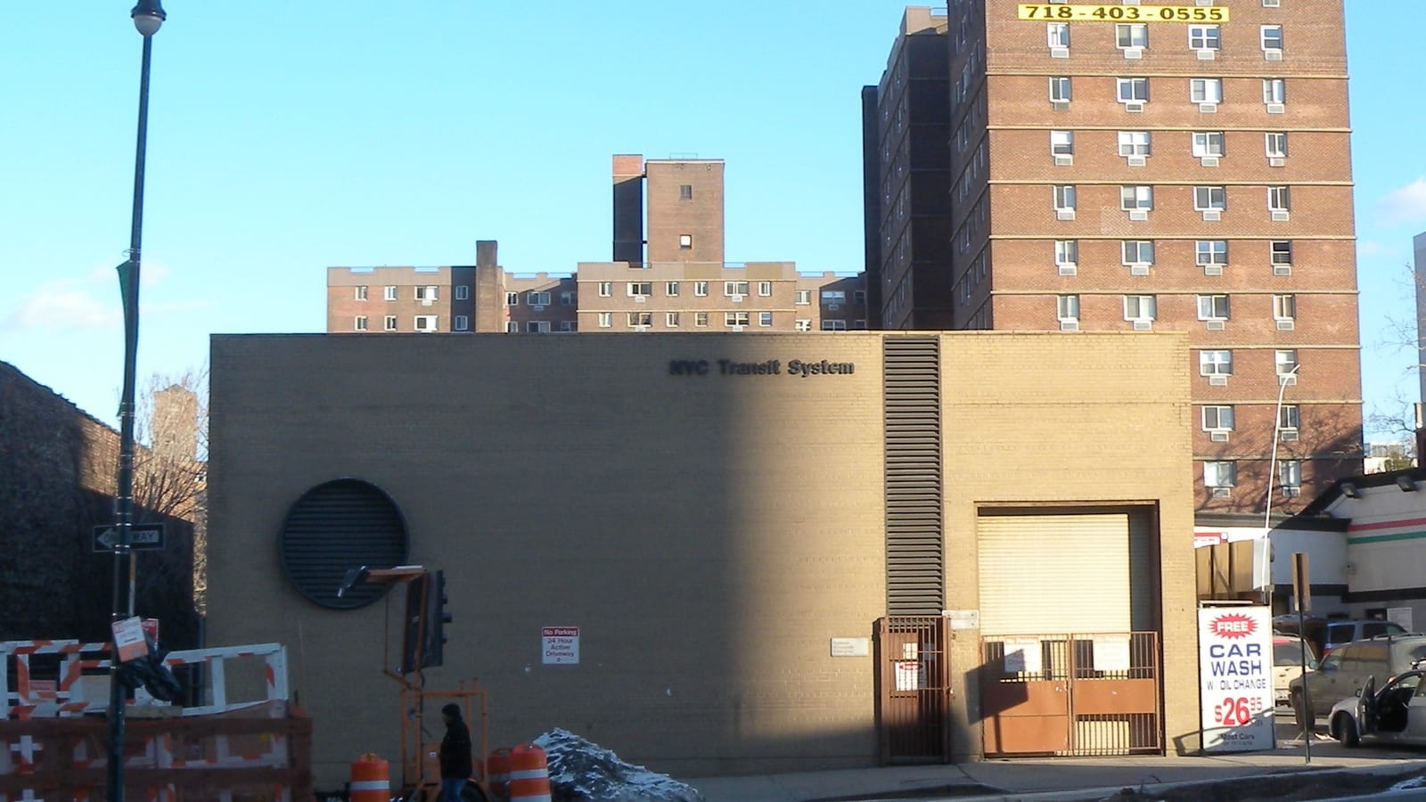 Photograph of a low tan brick building marked NYC Transit System with a large round vent and a roll-down gate, orange traffic barrels and a person walking in front, a car wash sign at right, and tall brick apartment buildings behind, one with a yellow phone-number sign, under a clear blue sky
