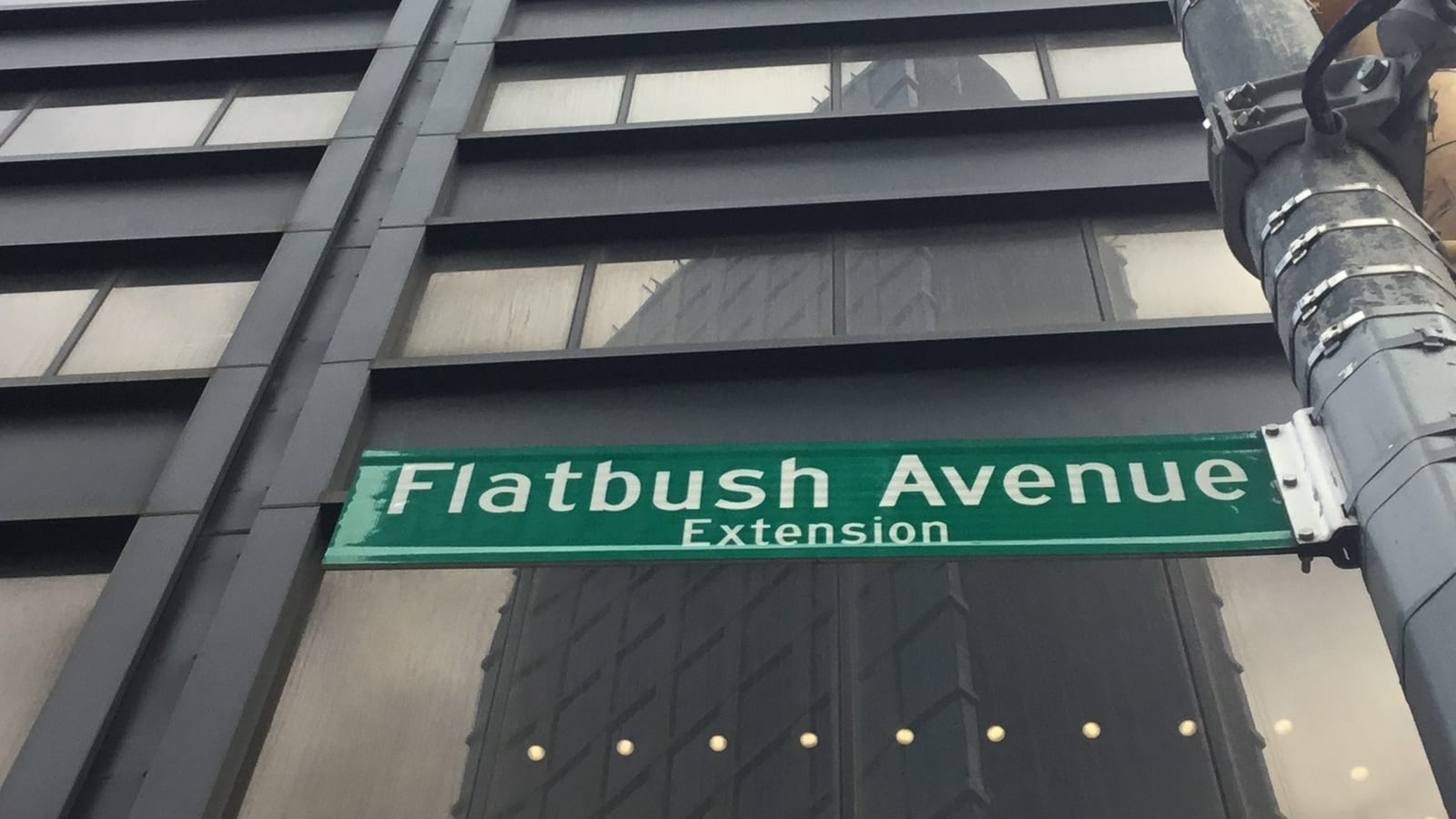 Photograph looking up at a green street sign reading Flatbush Avenue Extension on a gray metal pole, in front of a dark office building with bands of windows