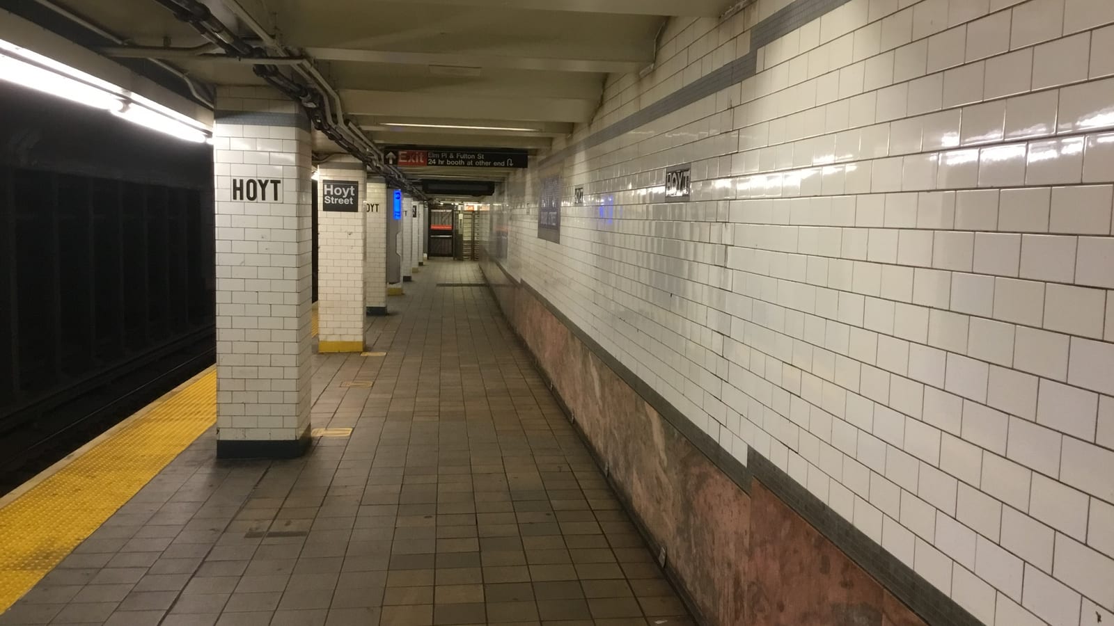 Photograph of an empty subway platform with white tiled columns marked Hoyt and Hoyt Street, a yellow platform edge at left, a pink marble wall base at right and an exit sign for Elm Pl and Fulton St in the distance