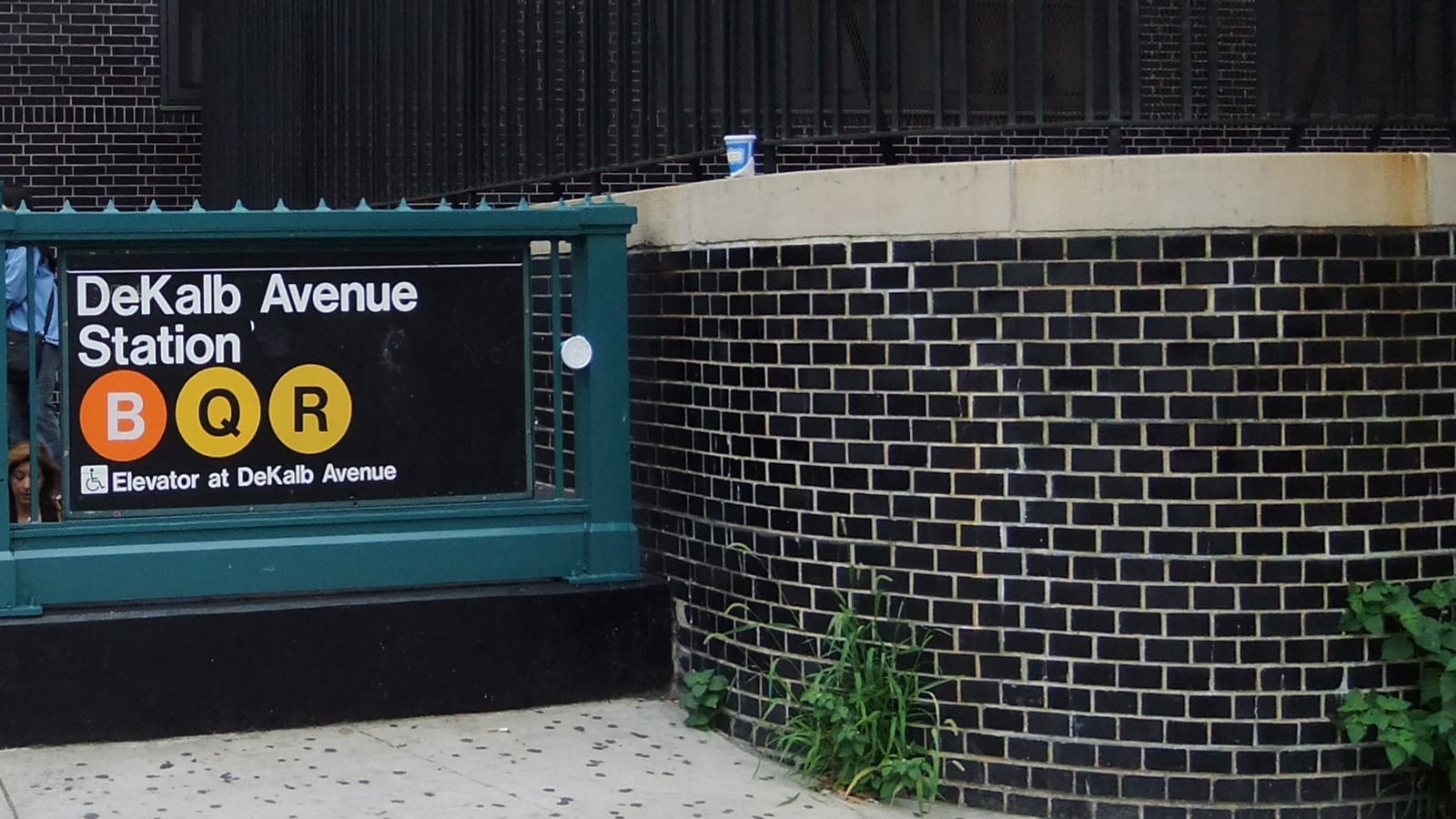 Photograph of a green iron subway entrance with a black sign reading DeKalb Avenue Station, round B, Q and R line symbols and Elevator at DeKalb Avenue, beside a curved wall of dark glazed brick with weeds at its base and a tall black iron fence above