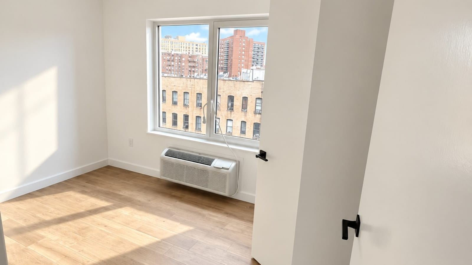 Empty room with a large window looking out on brick apartment buildings and a wall heating and cooling unit under the window