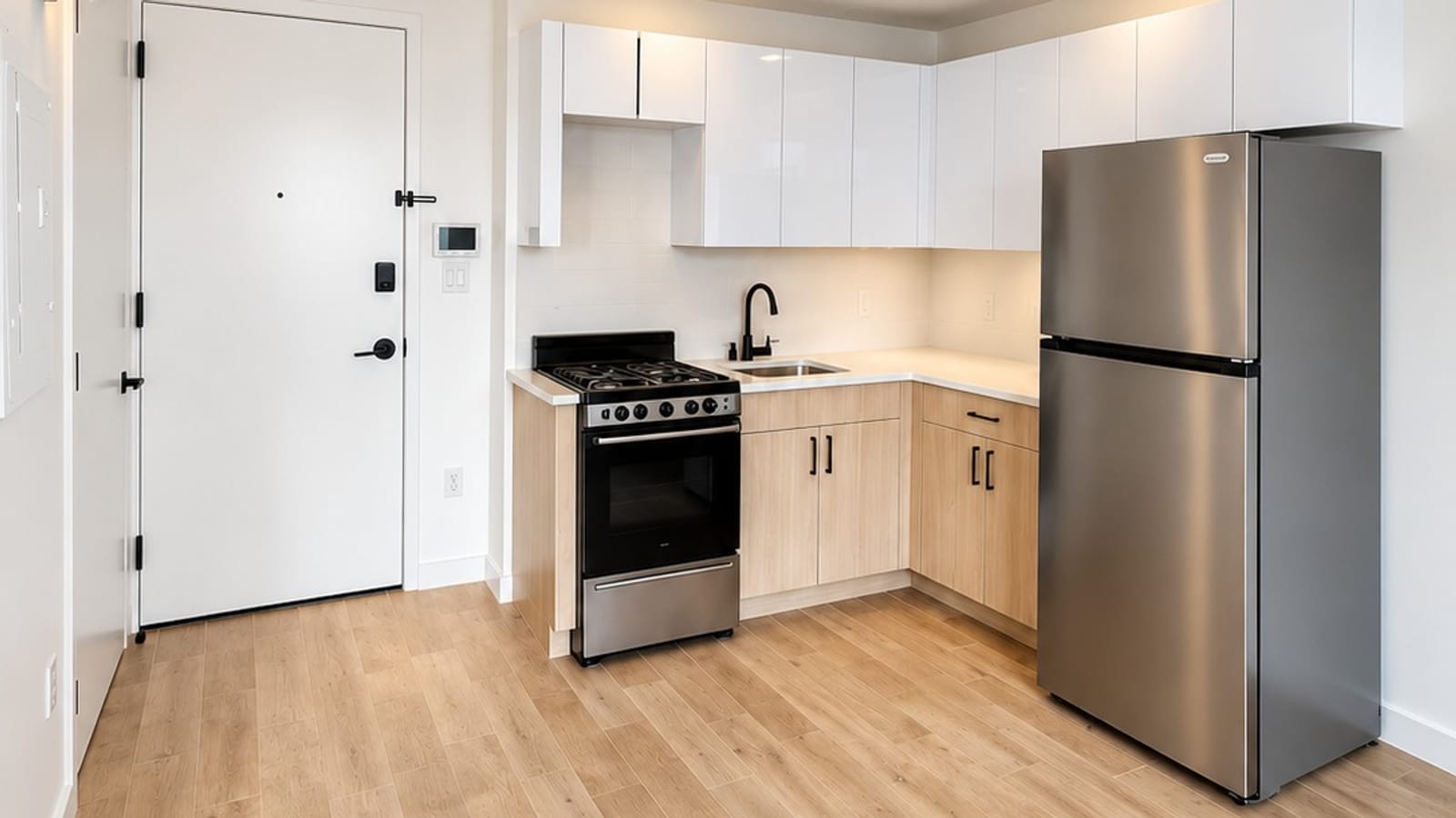 Kitchen with white upper cabinets, light wood lower cabinets, a stainless steel range and refrigerator, next to the apartment door